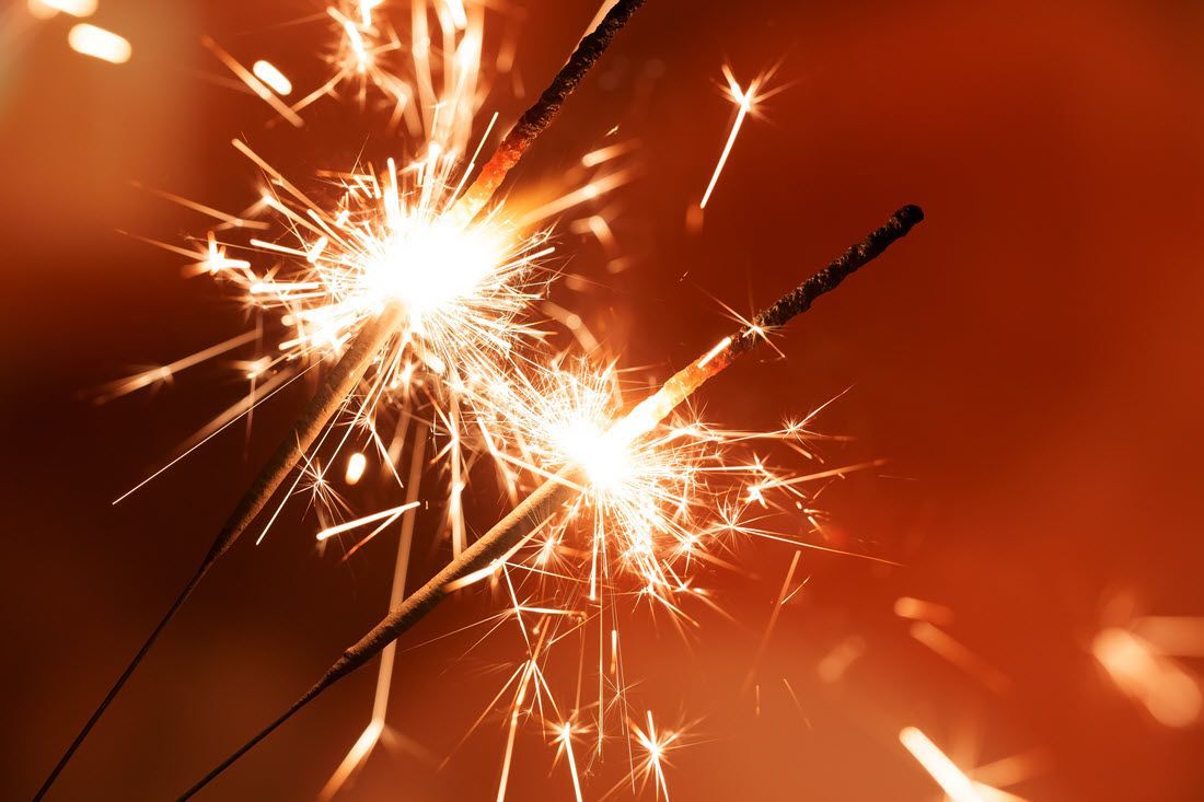 Two lit sparklers crossing against a warm orange background, sending bright sparks outward