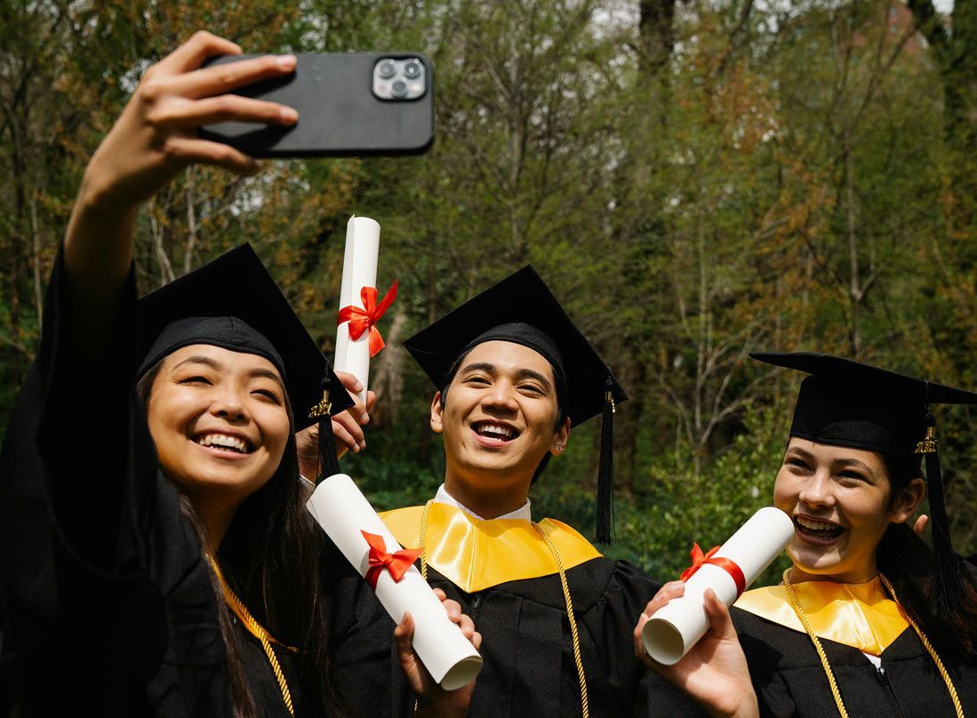 Three graduates in caps and gowns smile while holding diplomas and taking a selfie outdoors