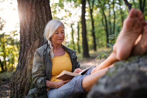 Woman reading a book in a forest, leaning against a tree with her bare feet up.