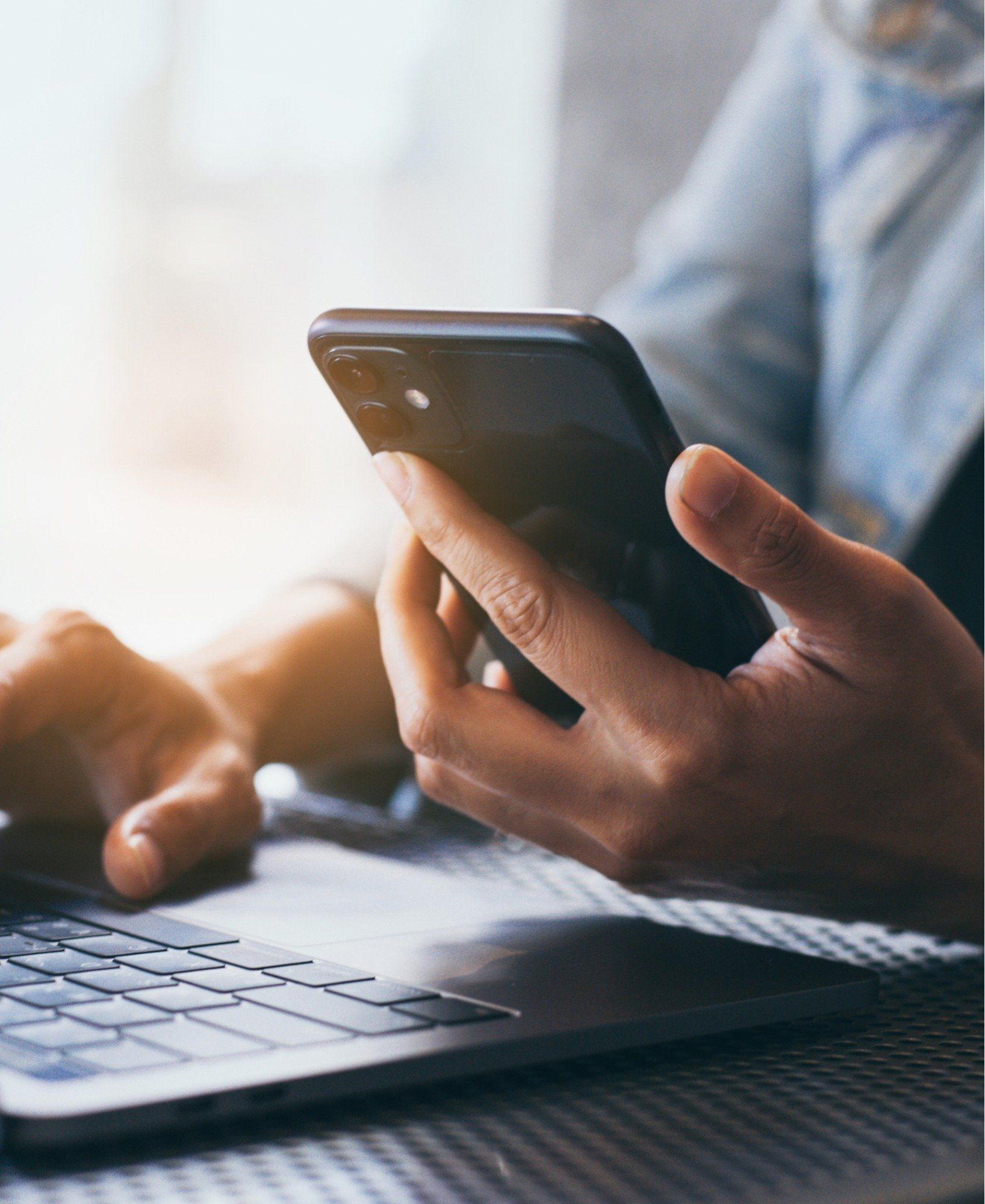 A person is typing on a laptop while holding a cell phone.