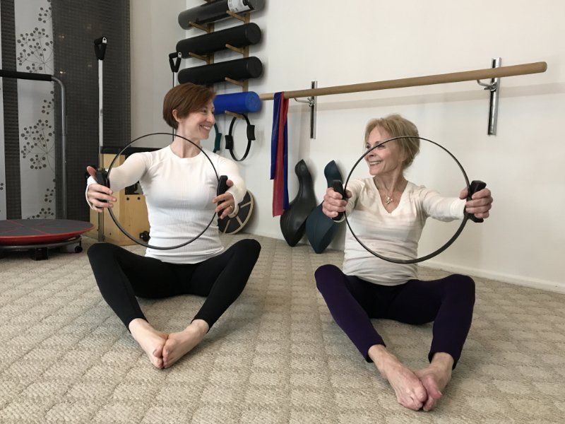 Two women are sitting on the floor holding Pilates magic circles.