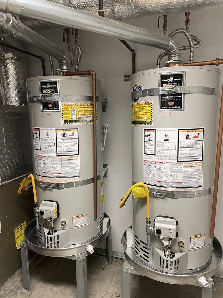 Two upright water heaters in a utility room. Gray tanks with yellow gas lines, silver metal stands, and exposed copper pipes.