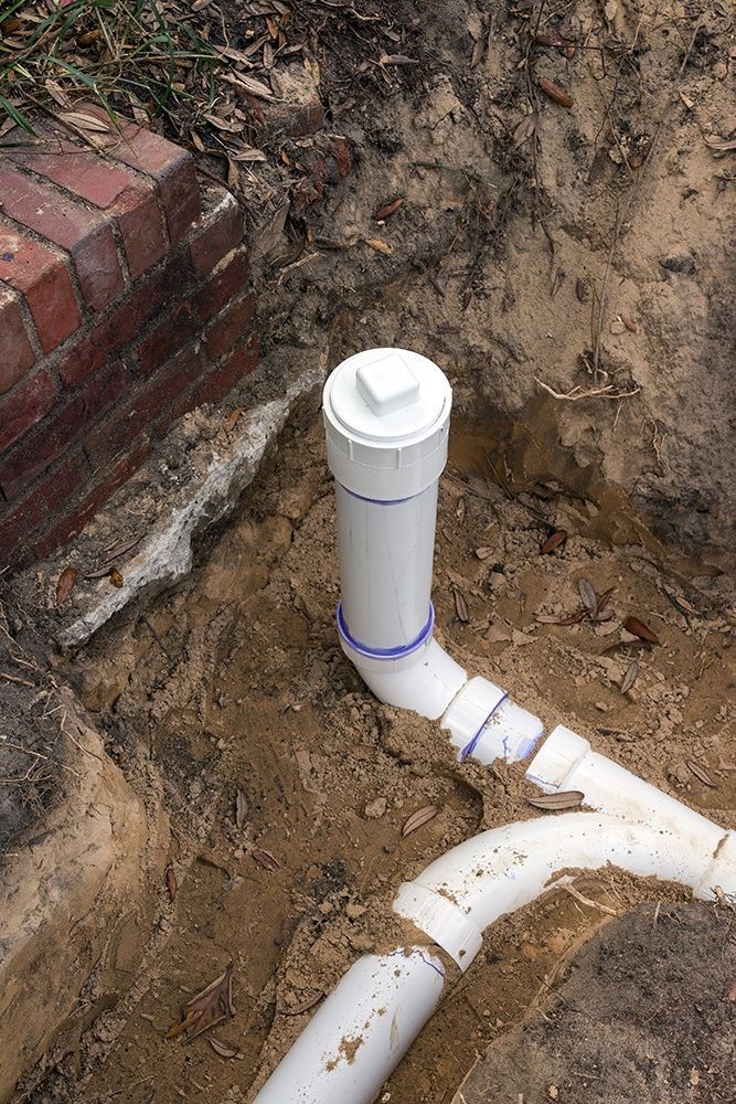 White PVC pipes in a dirt trench, connected to a brick wall.
