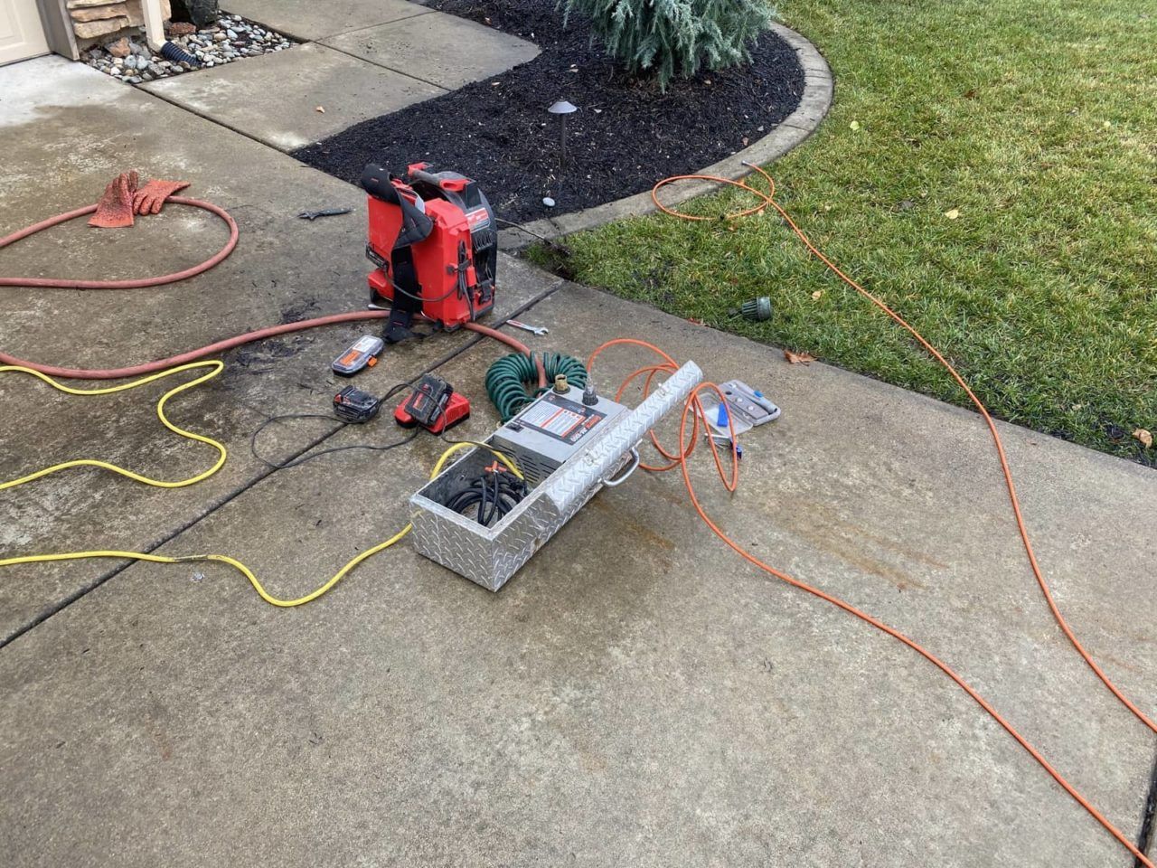Red generator, tools, and box on concrete driveway. Power cords and grass visible.