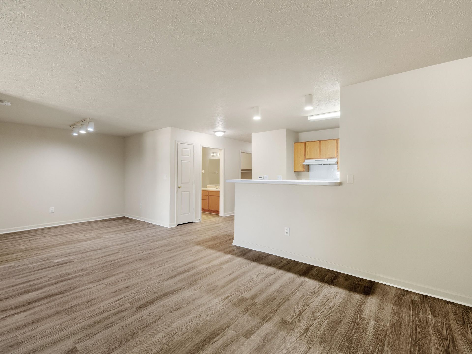 An empty living room with hardwood floors and a kitchen in the background.