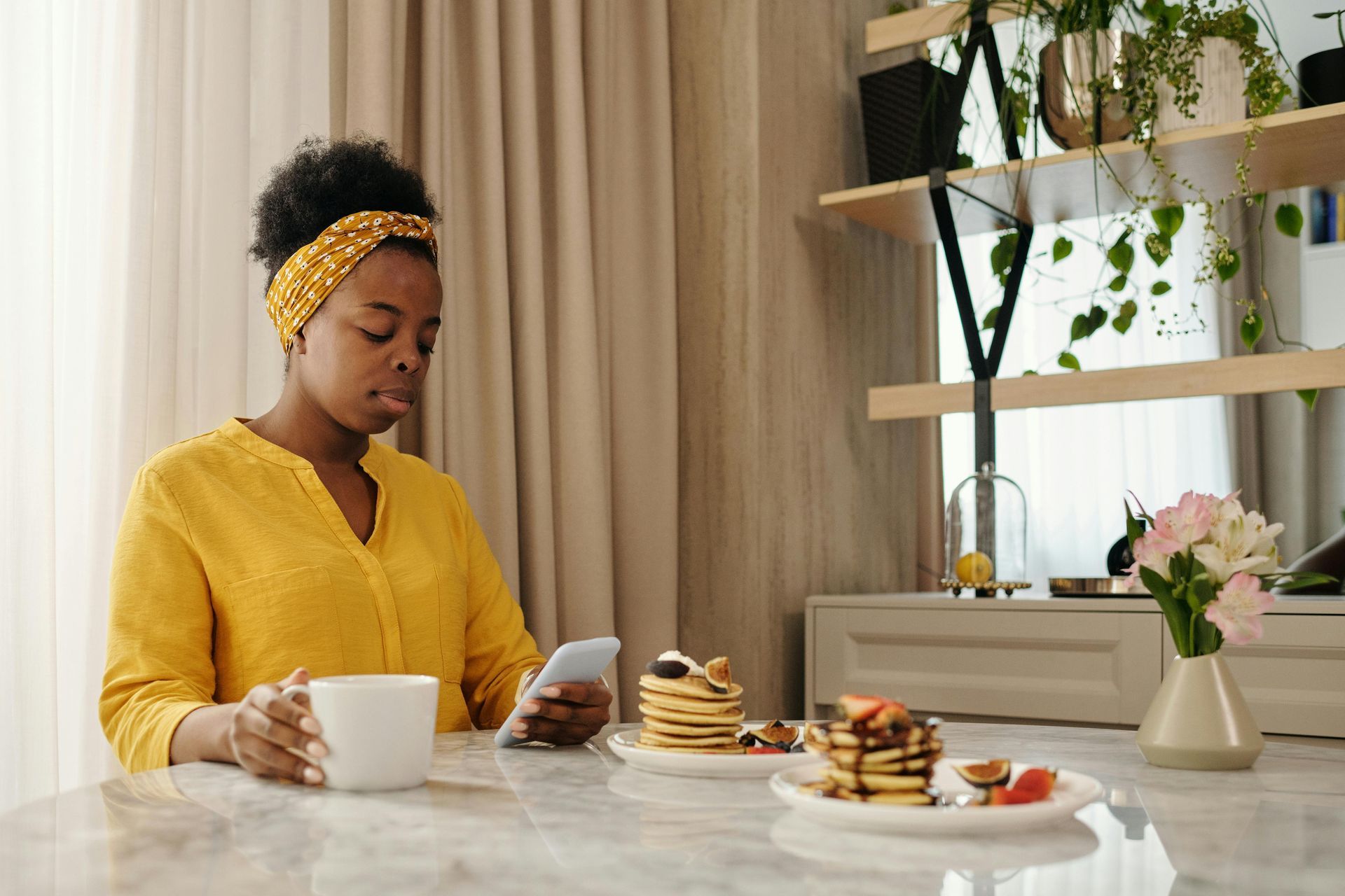 Vrouw in een geel shirt, aan het bellen, koffie drinkend en pannenkoeken etend aan een tafel in een goed verlichte ruimte.