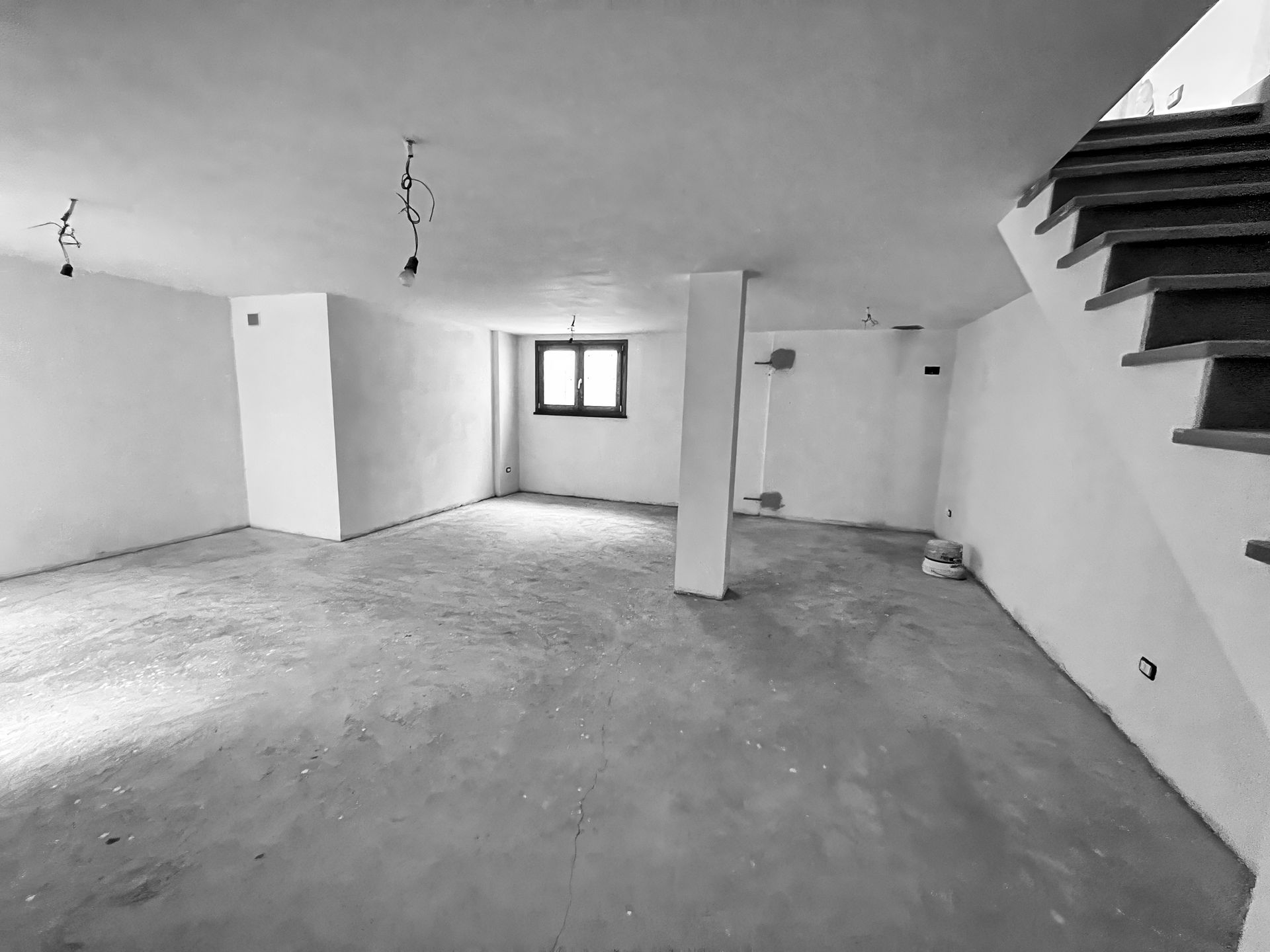 Empty, unfinished basement with concrete floor, white walls, support column, and stairs.