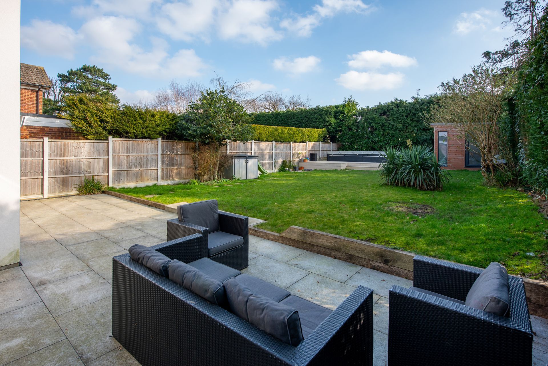 Patio with dark gray outdoor furniture, leading to a green lawn surrounded by hedges and a wooden fence.