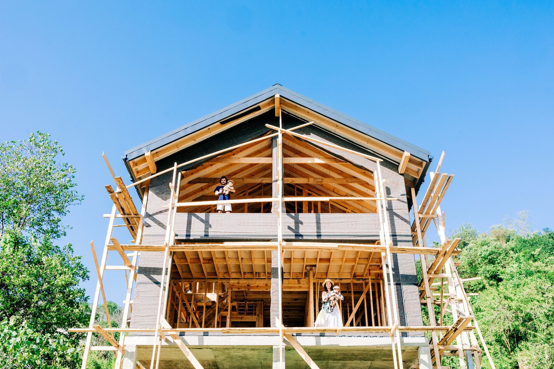 Couple in formal attire pose in an unfinished house under construction, framed by scaffolding, with blue sky.