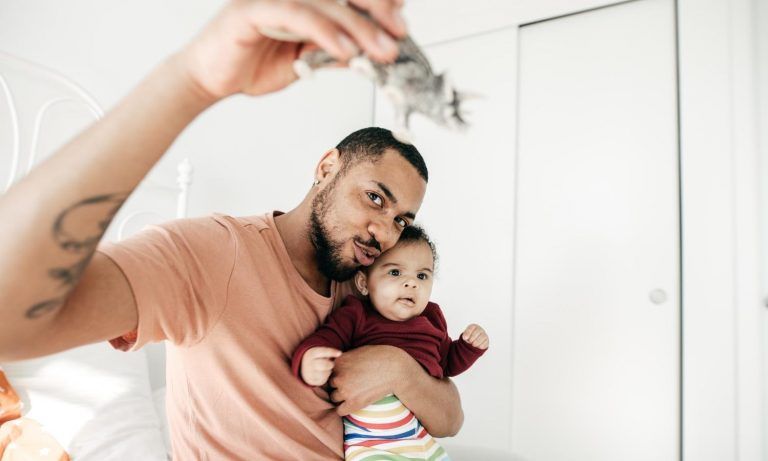 Man holding baby, showing toy. Both looking upward. Bedroom setting, neutral tones.