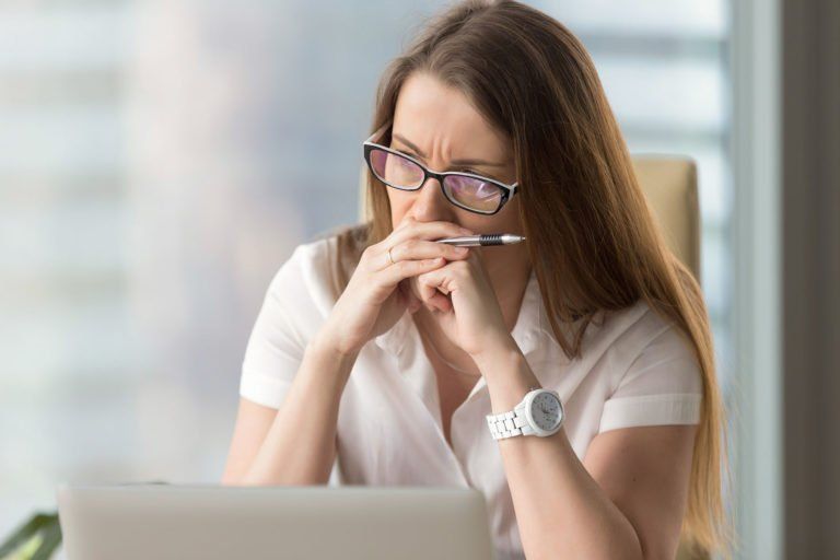 Woman wearing glasses, looking at a laptop with a furrowed brow, holding a pen, and resting her chin on her hands.