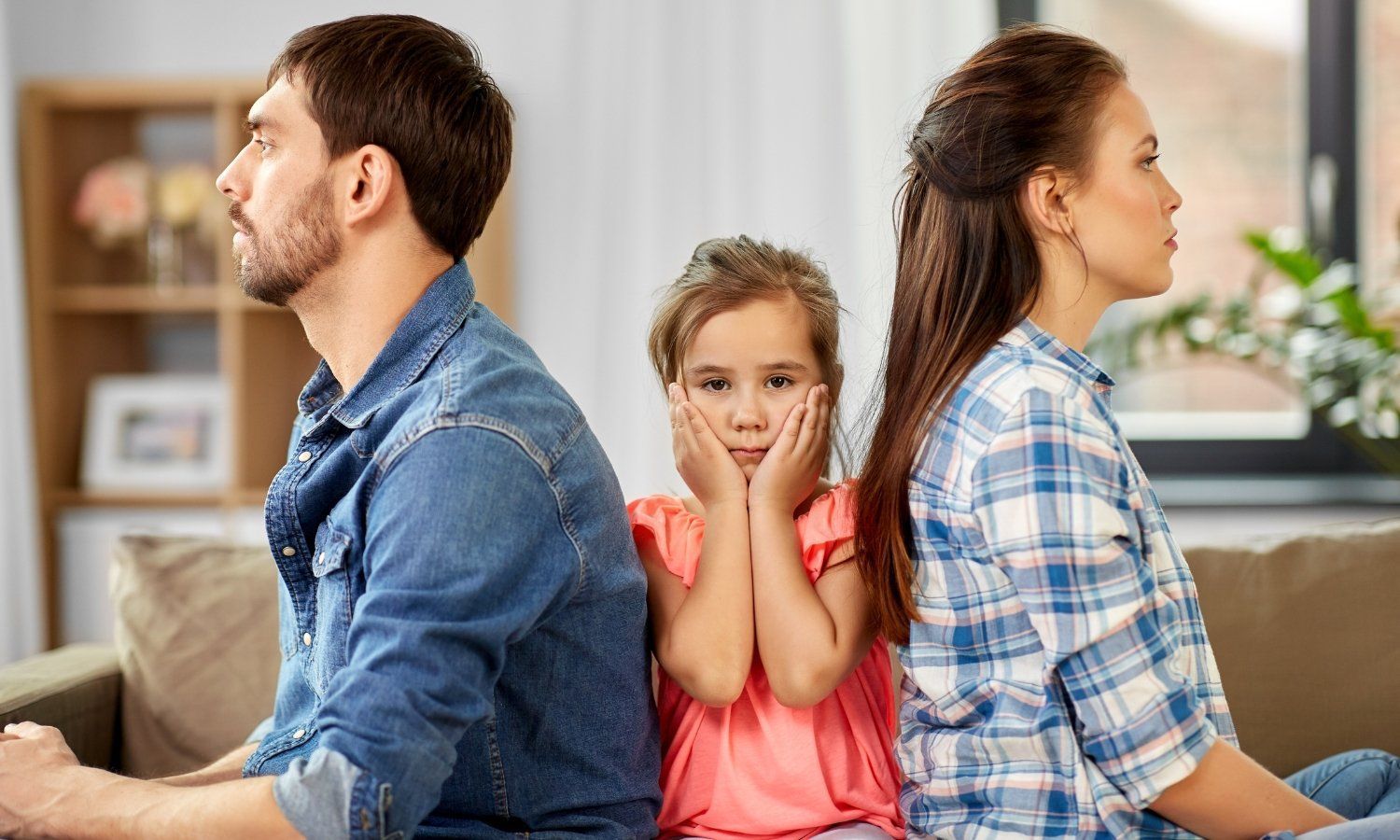 A girl looks concerned as parents sit apart, backs turned. Living room setting.