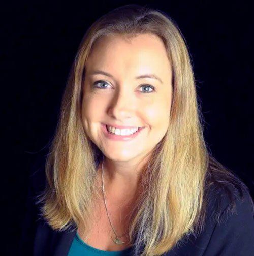 Woman with blonde hair smiling, wearing a teal top and black blazer; black background.