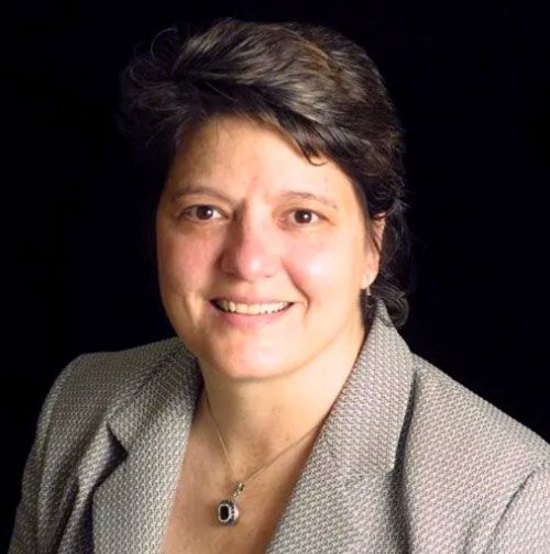 Woman with short dark hair smiling, wearing a gray blazer and necklace, against a black background.