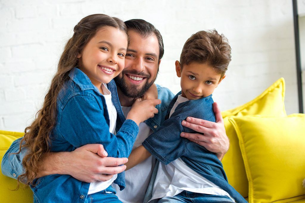 Man hugging two children on a yellow couch; all smiling.