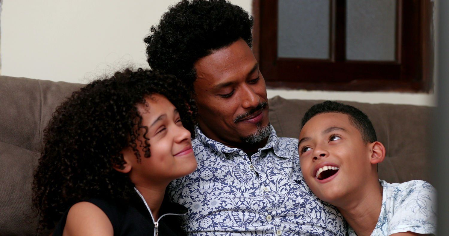 A father and two children smile, sitting close together on a couch.