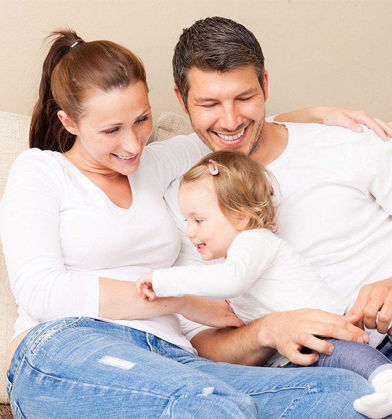 Family of three smiling, cuddling on a couch, indoors. Mom, dad, toddler daughter; all wearing white tops, jeans.