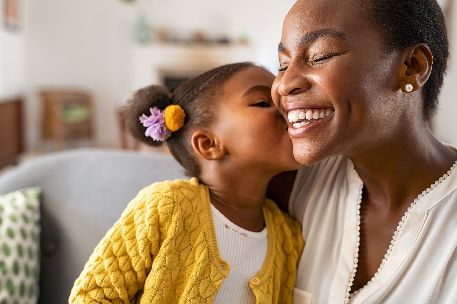 Young child kisses smiling woman's cheek. Child wears yellow sweater, woman has earring.
