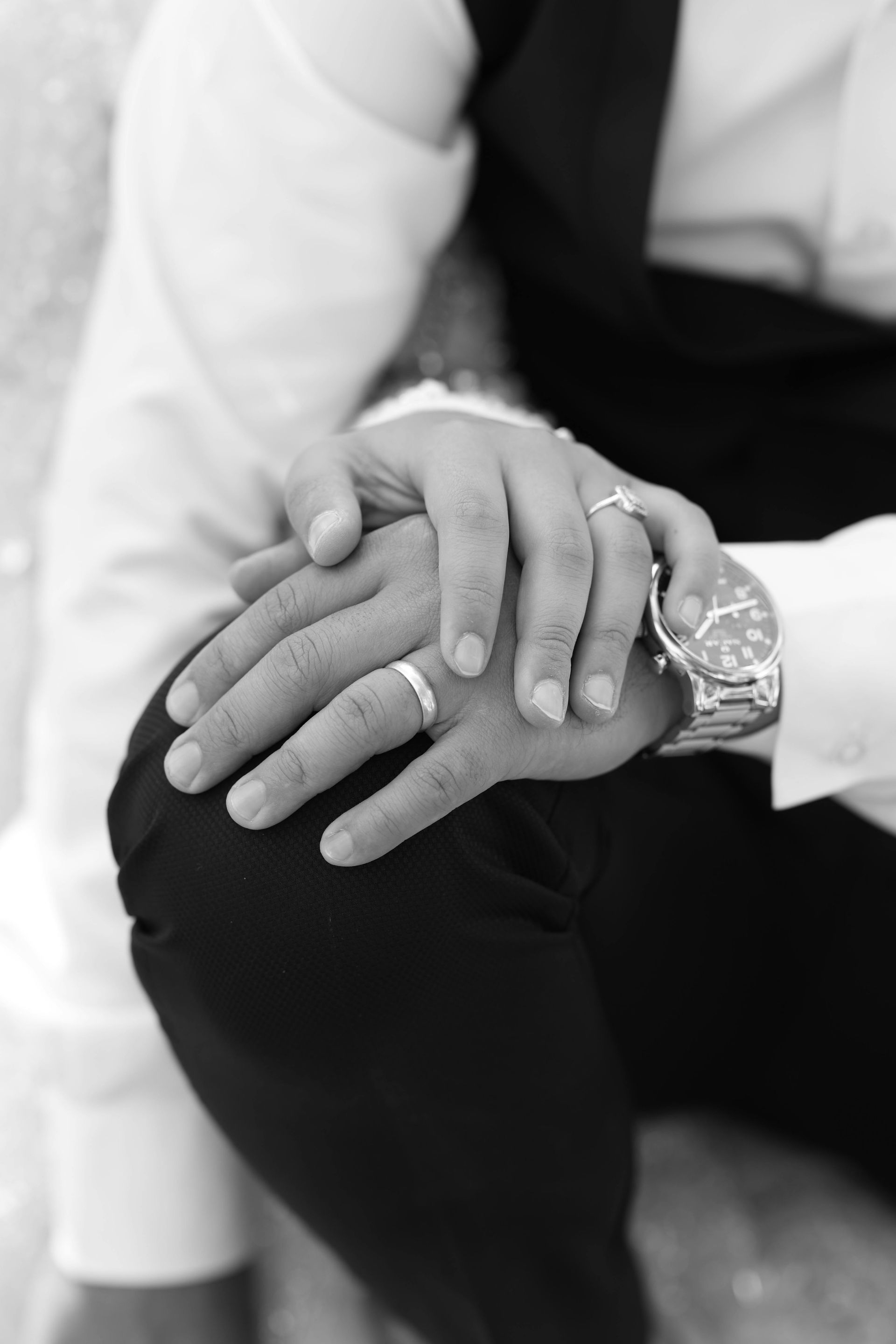 Two hands clasped, wearing wedding rings, one with a wristwatch, on a person wearing a vest and dress shirt.