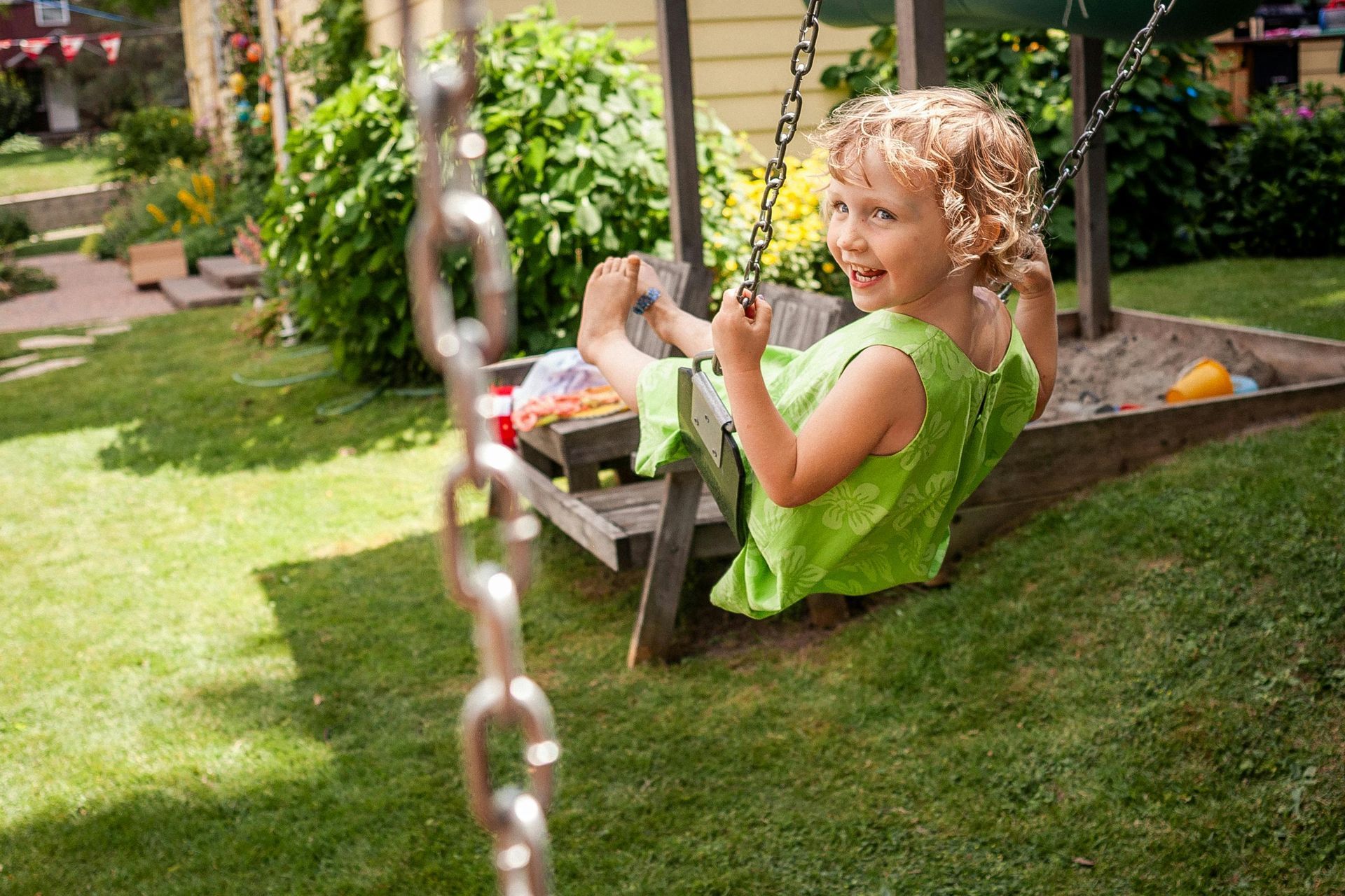 Child in green dress smiles, swinging on a wooden swing in a grassy backyard.