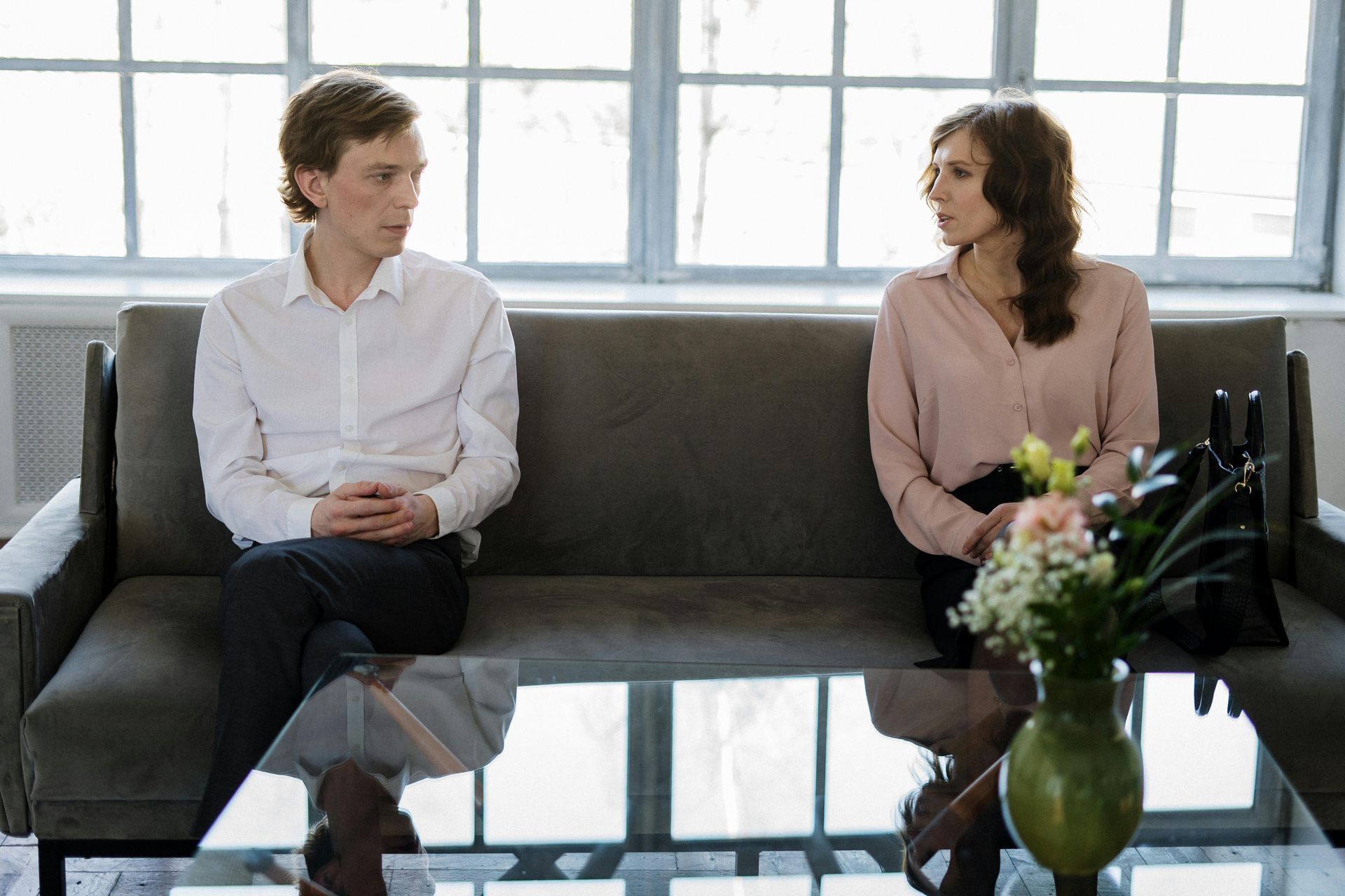 Man and woman seated on a sofa, facing each other. Interior setting, natural light. Serious expressions, talking.