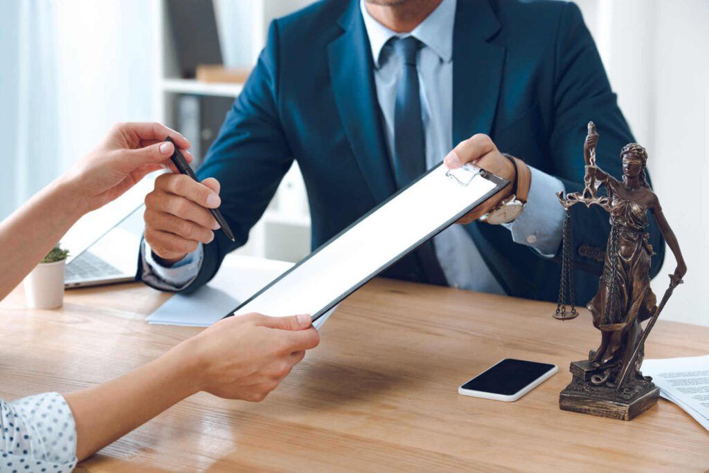 Person signing document with lawyer in office. Statue of Justice on desk.