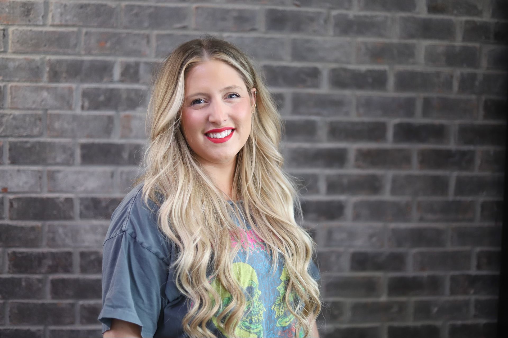 A woman with long blonde hair is smiling in front of a brick wall.