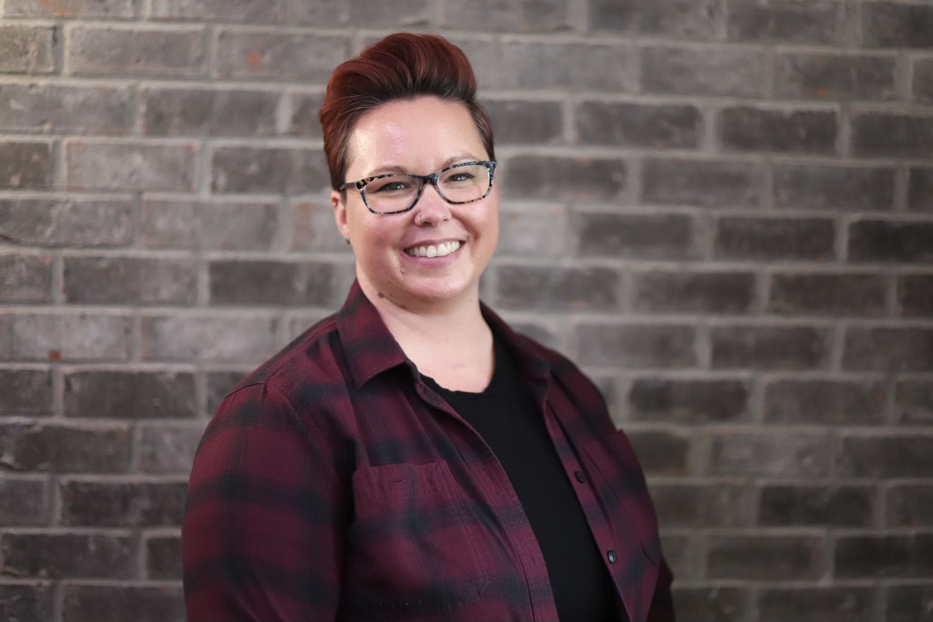 A woman wearing glasses and a plaid shirt is smiling in front of a brick wall.