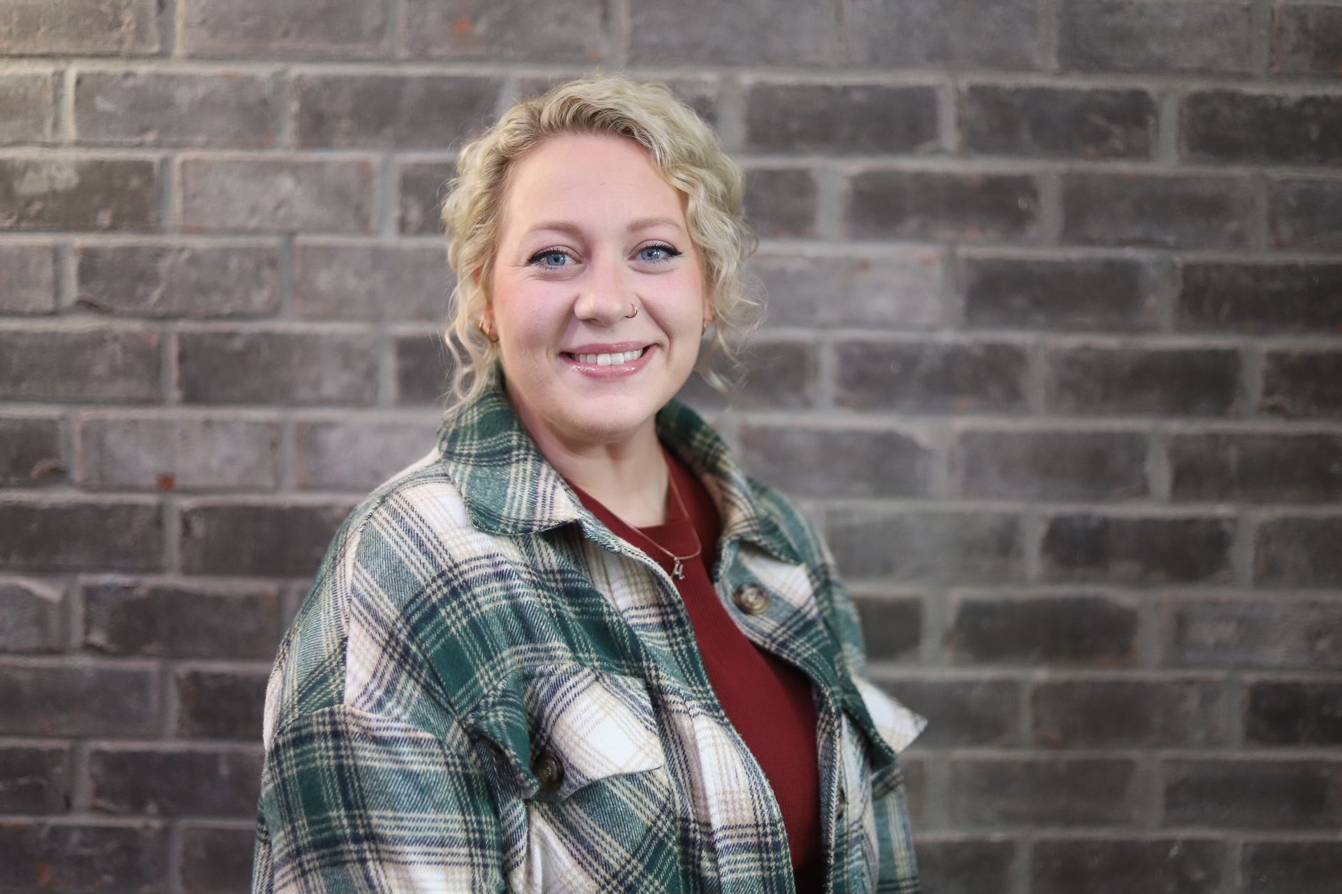 A woman in a plaid shirt is smiling in front of a brick wall.