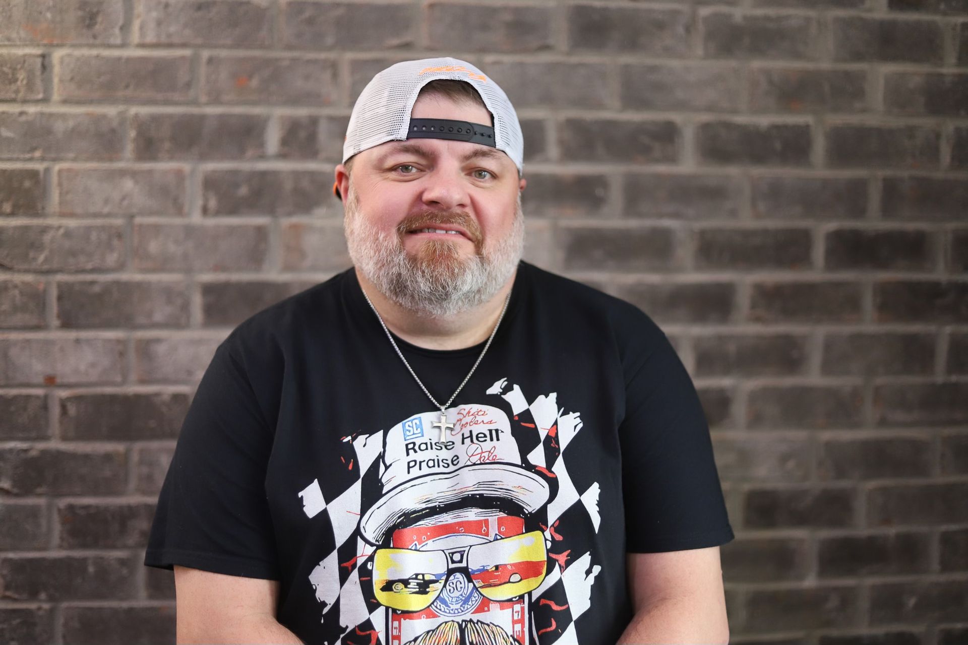 A man wearing a hat and sunglasses is standing in front of a brick wall.