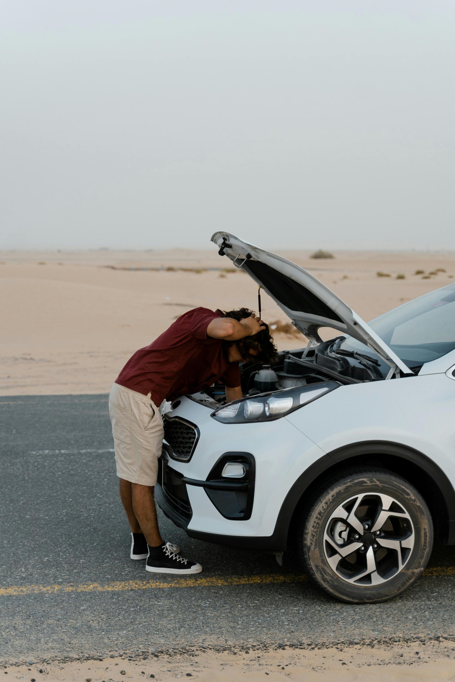 Man leaning into the open hood of a white SUV on a desert road, checking the engine.