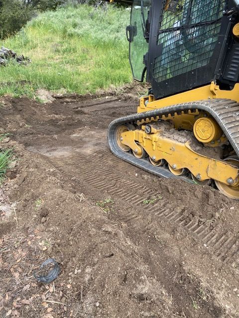 Yellow tracked skid steer moving dirt on a muddy path beside grass.