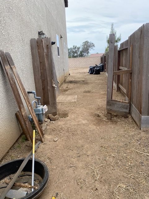 Narrow dirt alley beside a house and wooden fence, with pipes, a wheelbarrow, and yard tools along the wall.