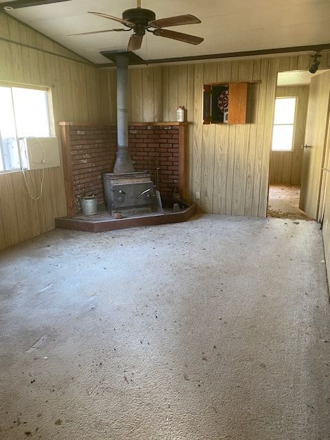 Empty room with wood stove, brick hearth, ceiling fan, and worn carpet in a small house