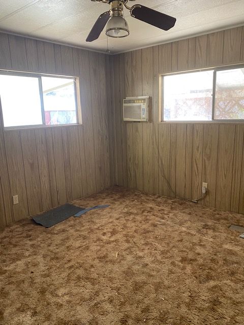 Empty wood-paneled room with patterned carpet, ceiling fan, two windows, and a wall air conditioner.
