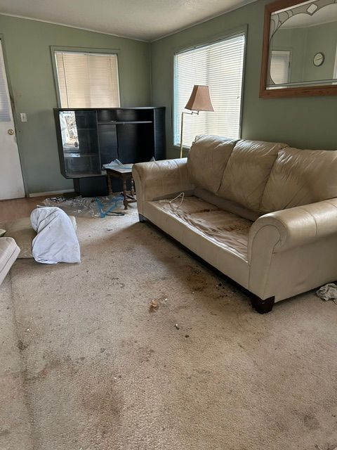 Messy living room with beige couch, black TV, lamp, and a white trash bag on stained carpet