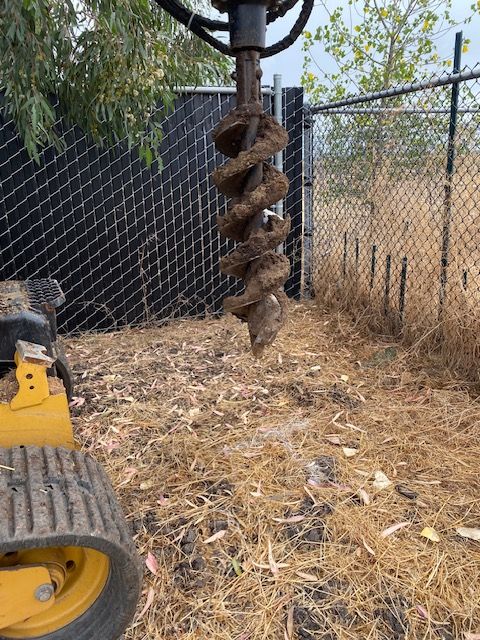 Large auger drill bit hanging over dry ground beside a chain-link fence and tractor wheel