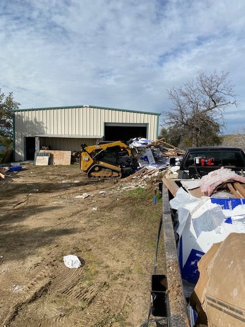 Damage-strewn yard beside a metal shed, with a yellow skid steer and piles of debris under a cloudy sky