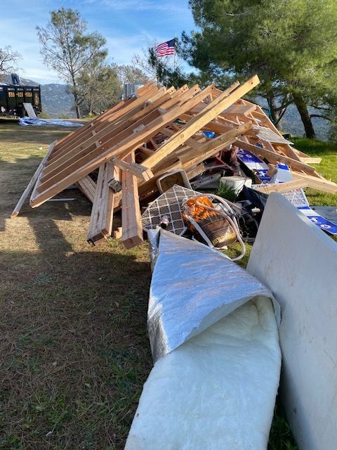 Collapsed wooden debris and torn white tarp piled on a grassy roadside near water under a sunny sky