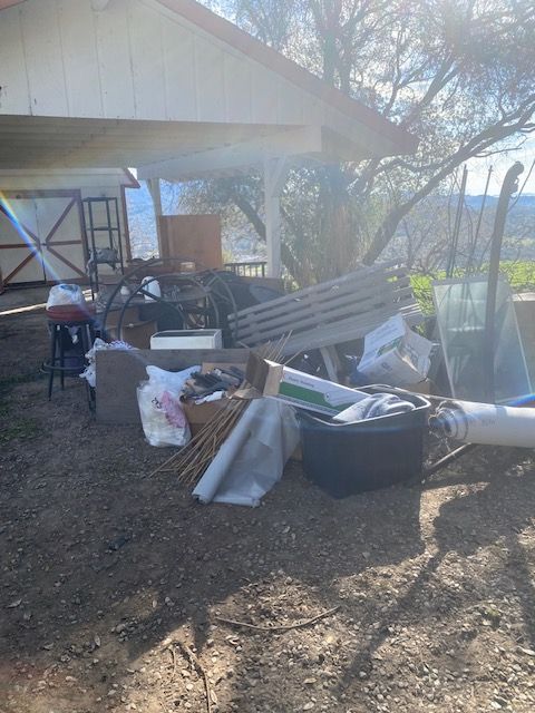 Outdoor cluttered yard with a shed, stacked materials, bins, and scattered debris in bright sunlight