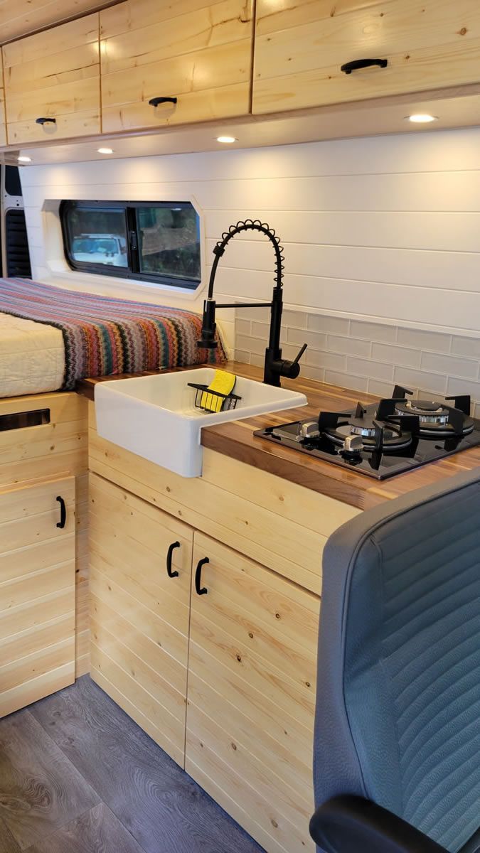 A kitchen in a van with a sink , stove and a bed.