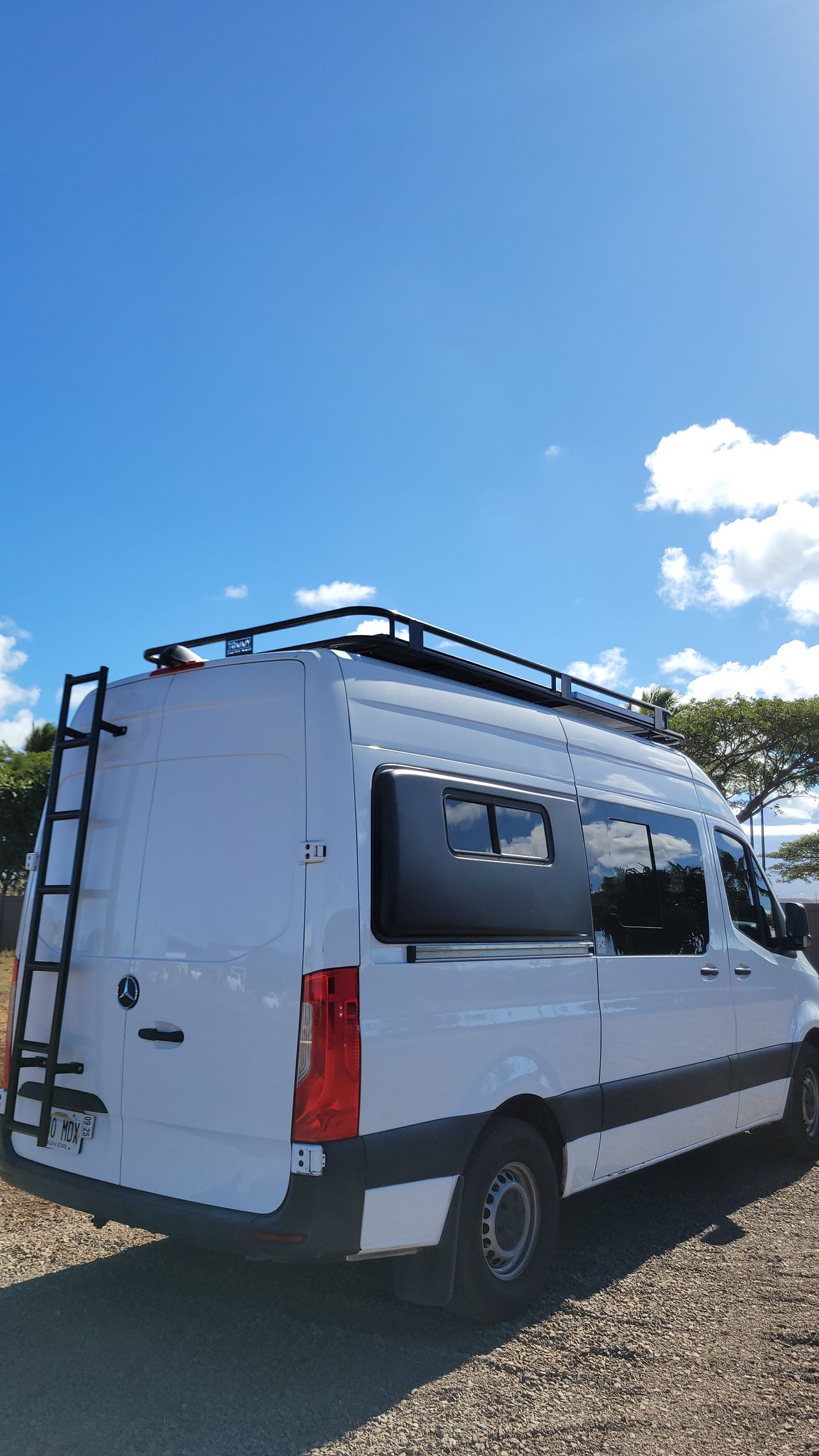 A white van with a ladder on the roof is parked in a gravel lot.