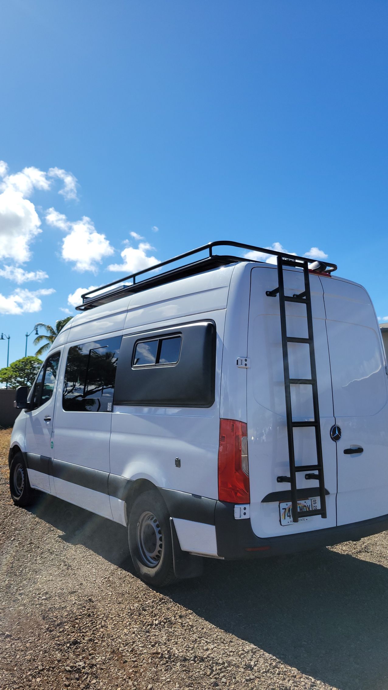 A white van with a ladder on the back is parked in a gravel lot.