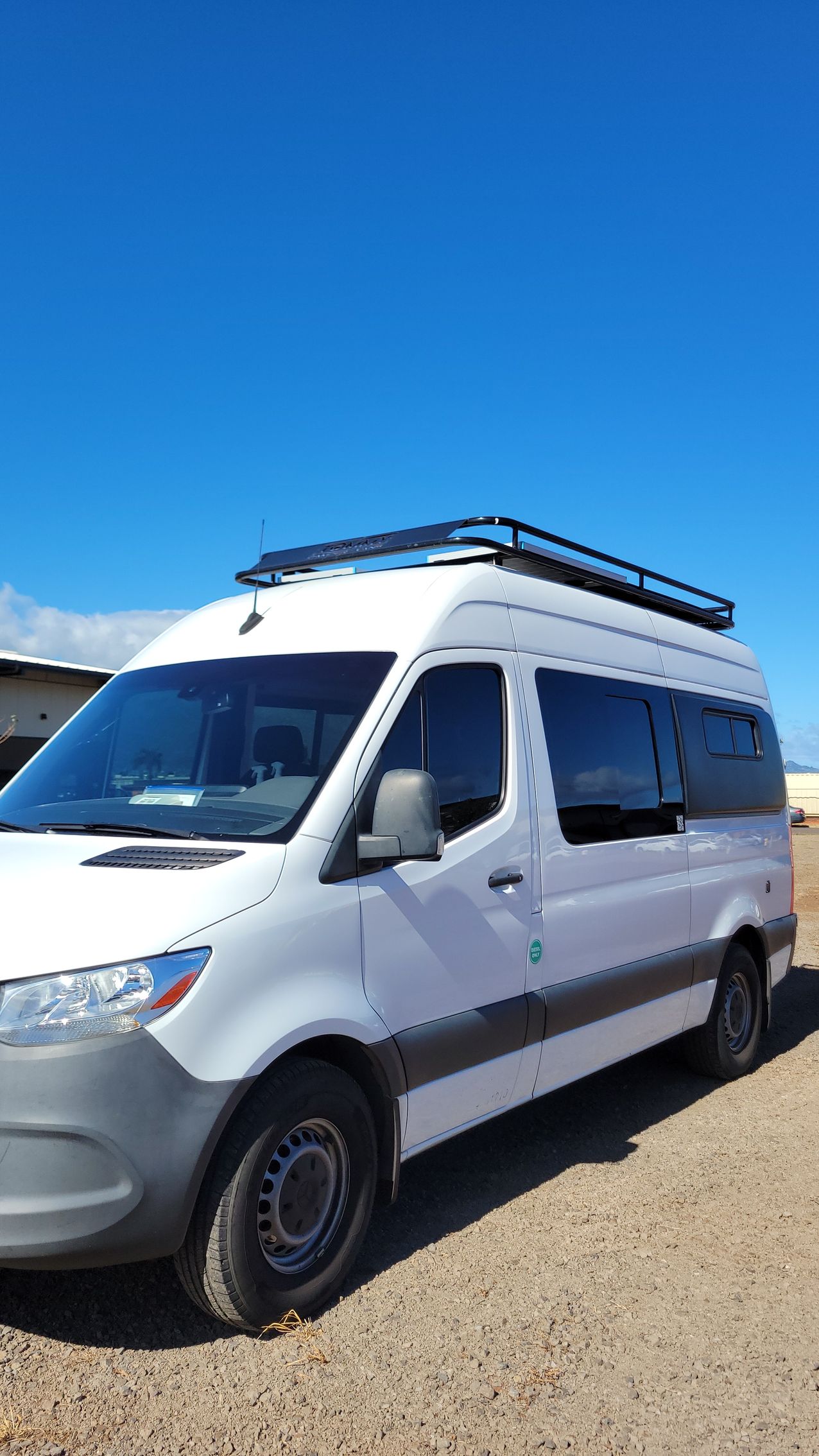 A white van with a roof rack is parked on a dirt road.