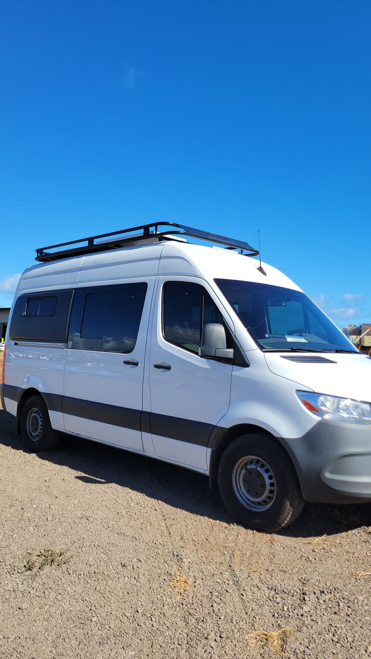 A white van with a roof rack is parked on a dirt road.