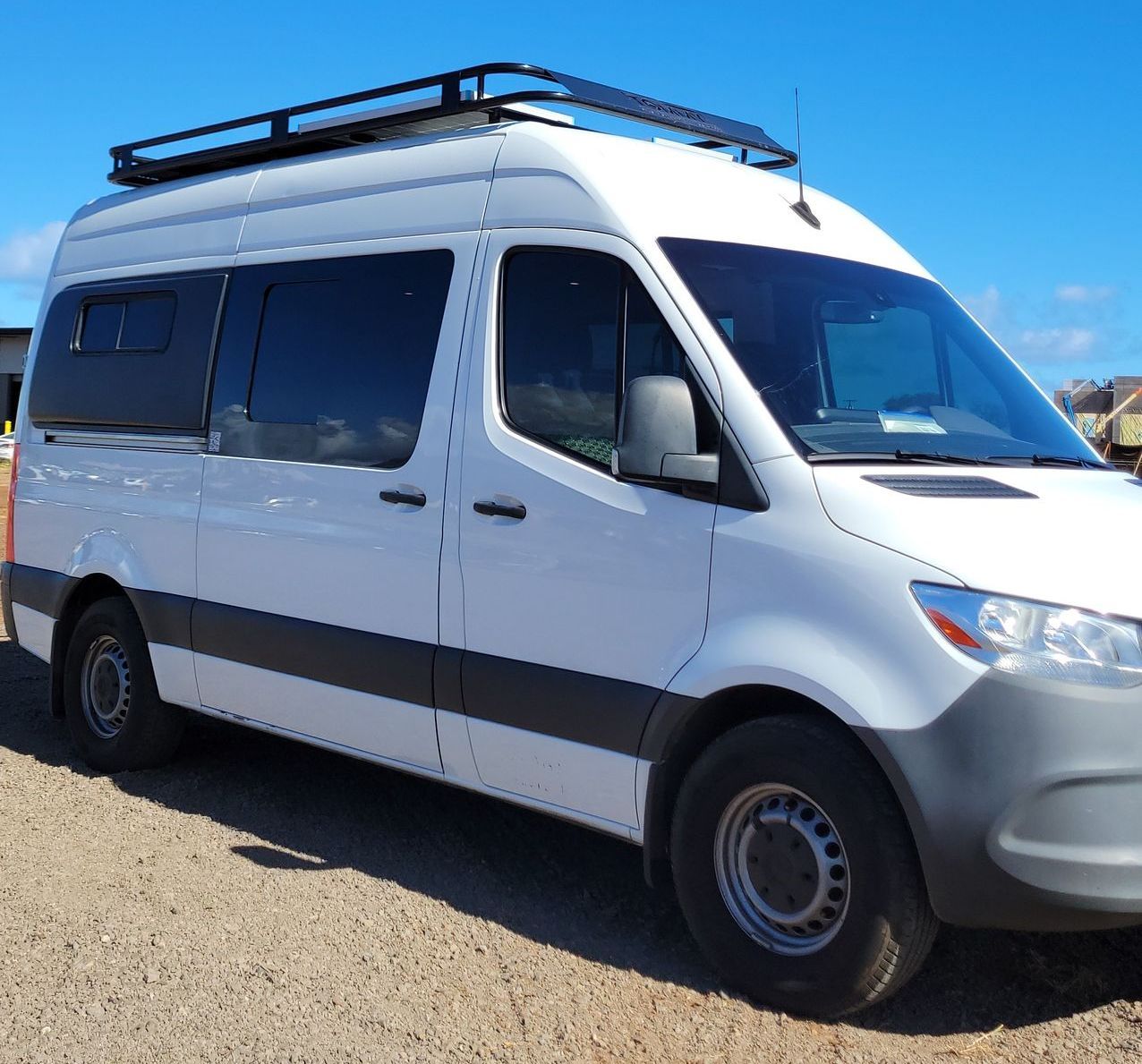 White camper van parked on a gravel lot under a clear blue sky, equipped with roof rack and tinted w