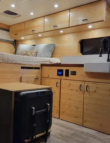 A camper van with wooden cabinets , a bed , a sink and a refrigerator.