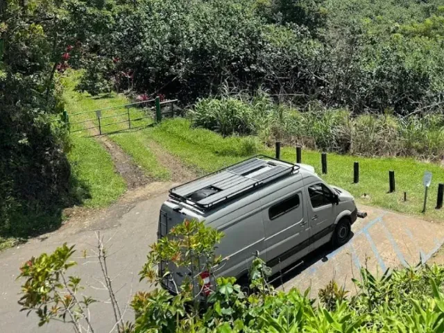 Gray campervan parked on a paved area with greenery, a gravel road, and trees in the background.