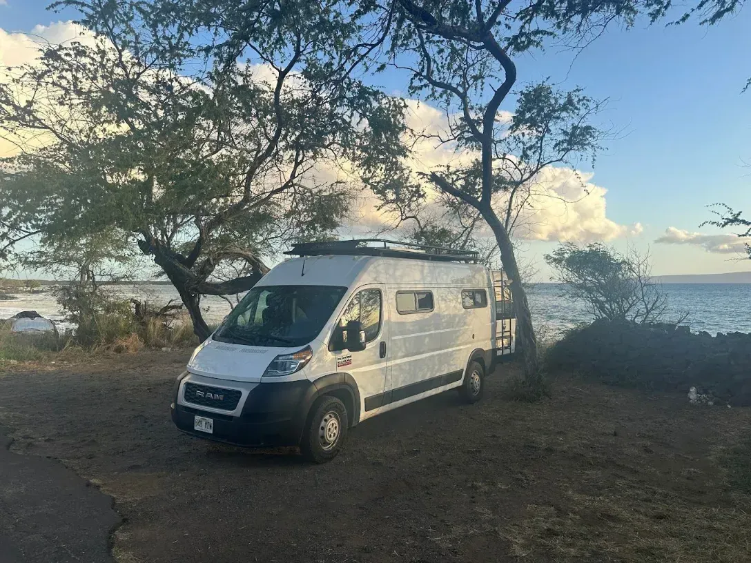 White camper van parked near a shore with trees. Blue water and sky in background.