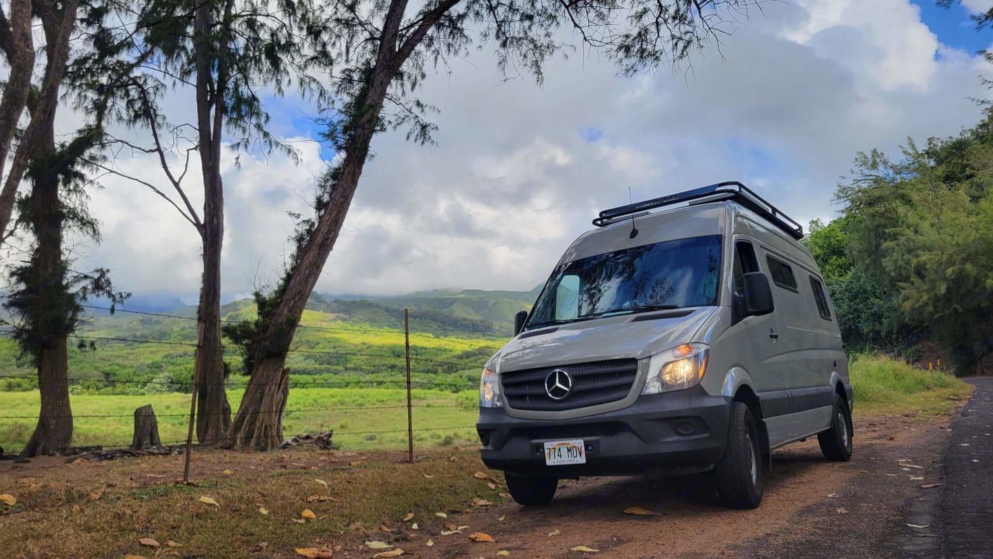 A Mercedes camper van from Mana Vans Hawaii parked roadside near Ke‘anae Uka, surrounded by lush green hills and cloudy skies. Tall trees frame the scene, highlighting the peaceful, remote feel of East Maui’s countryside.